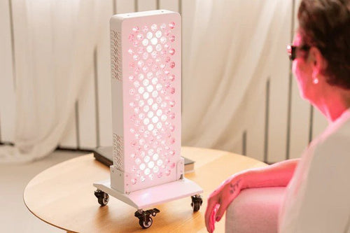 Vrouw met beschermbril zit naast een brandend rood licht therapie paneel op een houten tafel